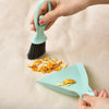 A hand is holding a teal mini broom and dustpan set on a beige surface.