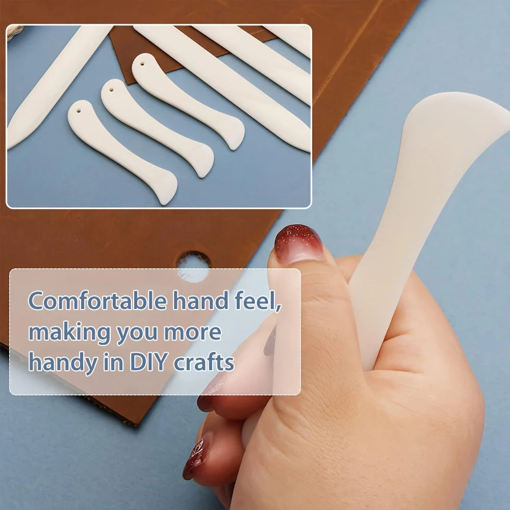 A hand holds a set of white plastic bone folders and crease markers for DIY crafts, shown next to a brown box.