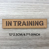 A brown woven patch with the text IN TRAINING is displayed on a wooden surface.