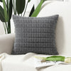 A gray knitted pillowcase with a textured corduroy pattern resting on a white sofa next to green leaves.