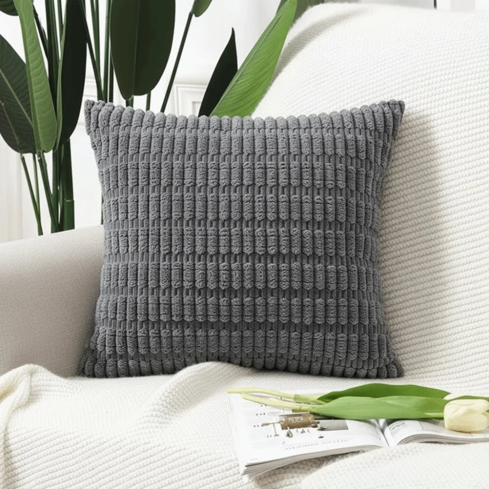 A gray knitted pillowcase with a textured corduroy pattern resting on a white sofa next to green leaves.