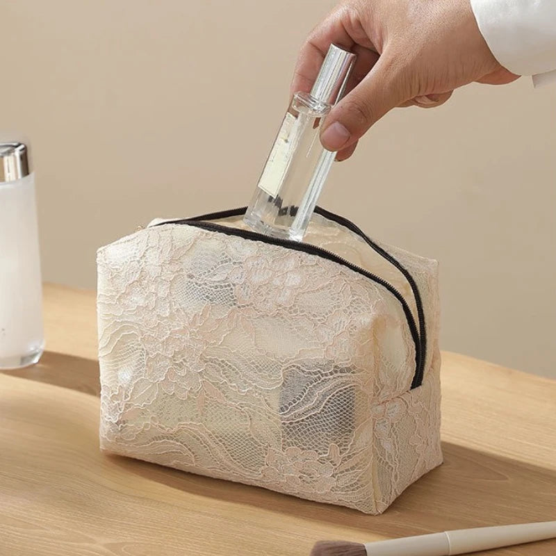 A hand is holding a clear glass bottle over a beige lace mesh makeup bag.