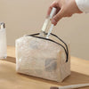 A hand is holding a clear glass bottle over a beige lace mesh makeup bag.