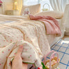 A hand is holding a light pink floral quilt blanket with a soft washed finish in a bright, airy bedroom setting.