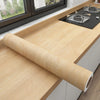 A roll of self-adhesive wood grain wallpaper film resting on a light wooden countertop.