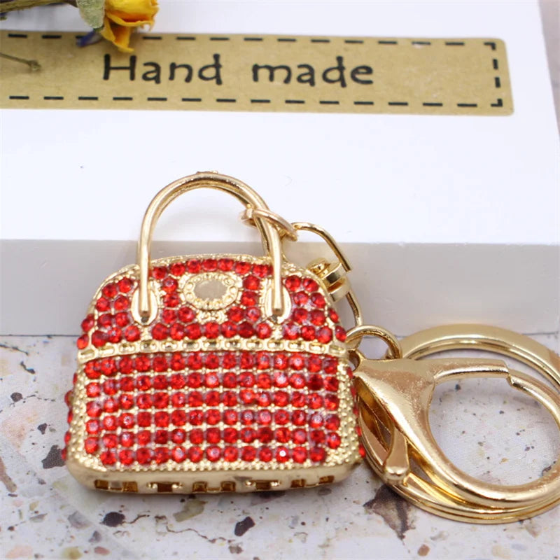 A small red crystal rhinestone handbag keychain with a gold-tone chain, displayed on a white box.