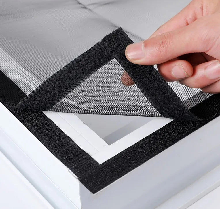 A hand is folding a black self-adhesive window mosquito net screen over a white frame.