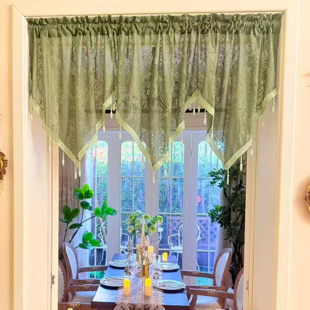 A sheer sage green lace curtain valance with fringe hangs over a window in a bright room.