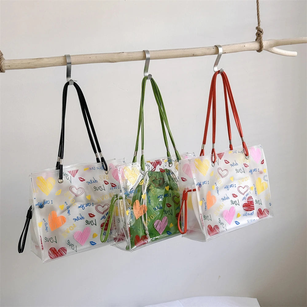 A set of transparent tote bags with inner pouches featuring pink and yellow hearts hanging from a wooden bar.