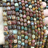 A hand holds a string of natural stone square faceted beads in various earthy and deep colors for jewelry making.