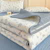 A stack of floral quilted bedspreads and a gray quilted cover with a matching fabric are displayed on a blue surface.