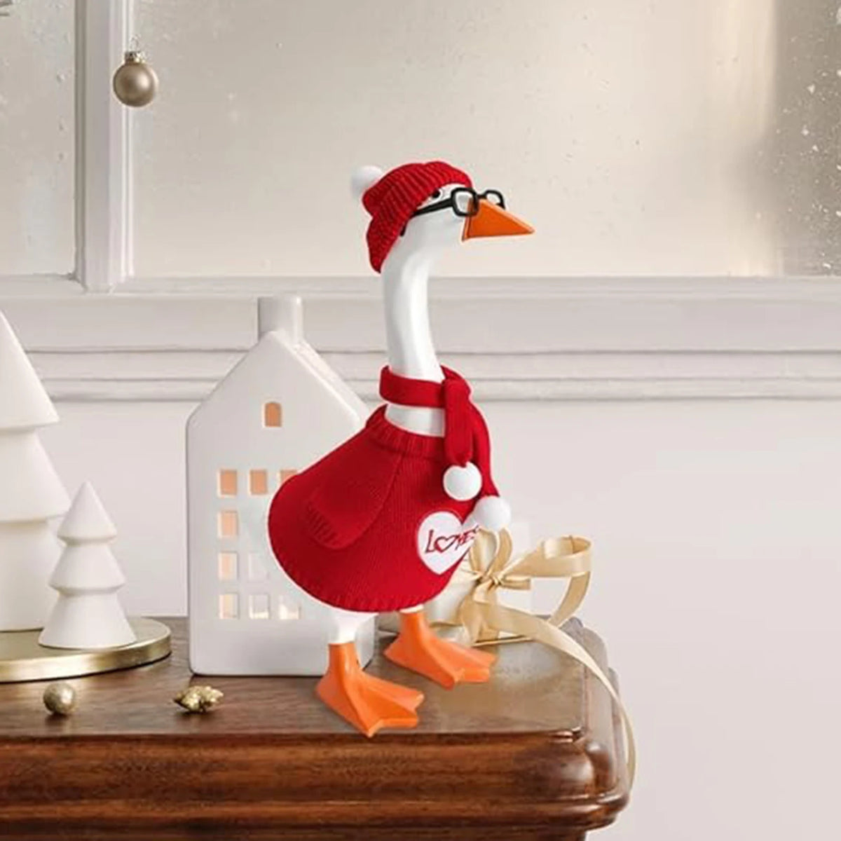 A white goose statue wearing a red knitted sweater, hat, and glasses, sitting on a wooden surface.