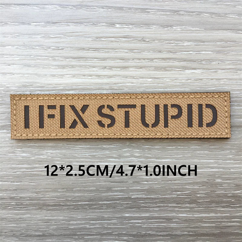 A rectangular brown woven patch with the text IFIXSTUPID printed on it, shown against a light wooden background.