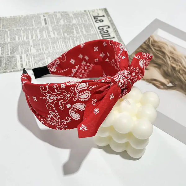 Diadema de seda roja con lazo estampado blanco junto a un producto de baño de burbujas blanco y un periódico.