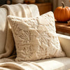 A cream-colored plush throw pillow with embossed maple leaf patterns resting on a light sofa.