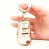 A hand holds a gold-toned protective TPU key fob cover with an oval shape and a circular loop.