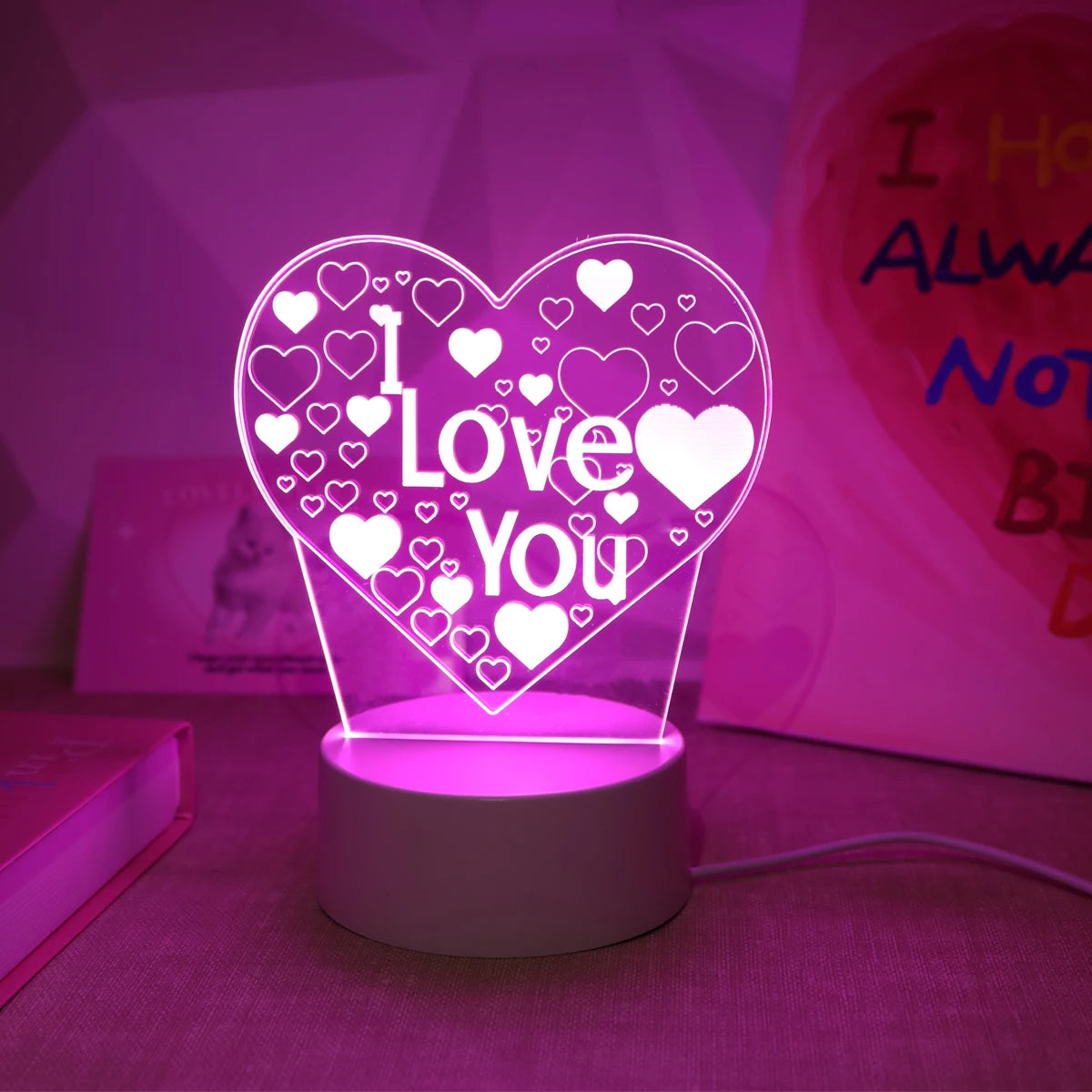 A glowing LED heart sign with 'I Love You' text in a 3D design sits on a stand against a pink background.