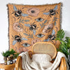 A bohemian jacquard woven throw blanket featuring floral and eye motifs, draped over a wooden chair.