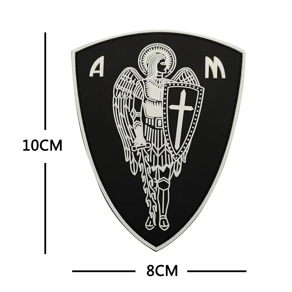 A black shield patch featuring a silver archangel St. Michael design with the letters AM.