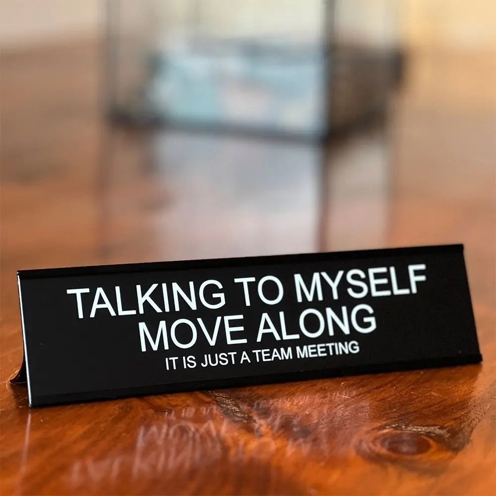 A black desk sign with white text that reads Talking to Myself Move Along It is just a team meeting