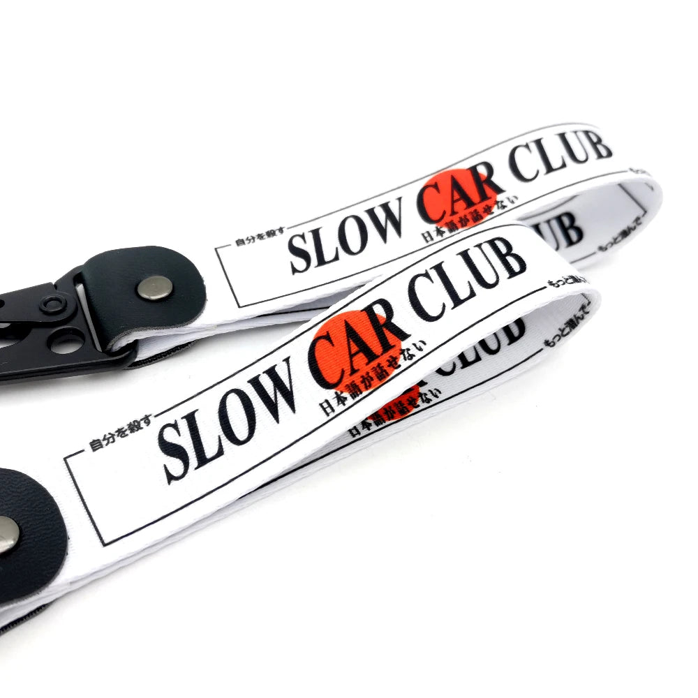A white nylon keychain with black steel clip featuring the text SLOW CAR CLUB in orange and black.