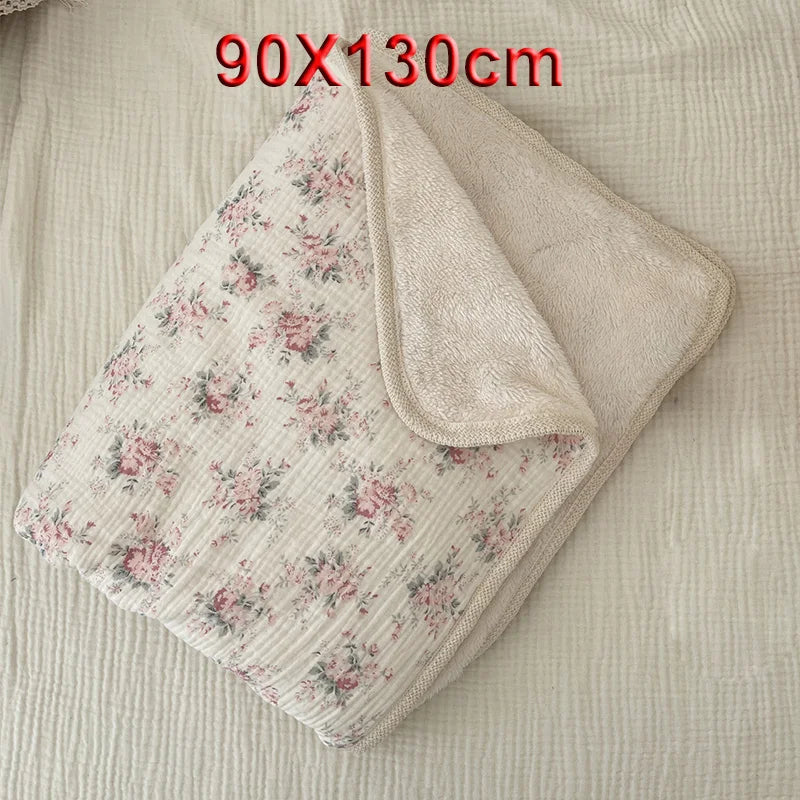 A light pink floral muslin baby blanket with a beige lining is shown folded on a light fabric background.