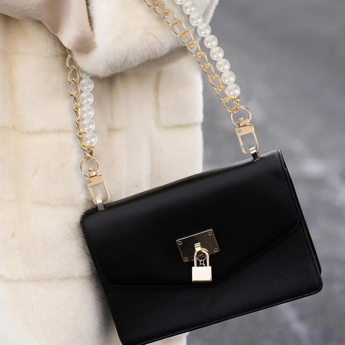 A black leather purse with a gold chain and pearl accents is shown against a white fur background.