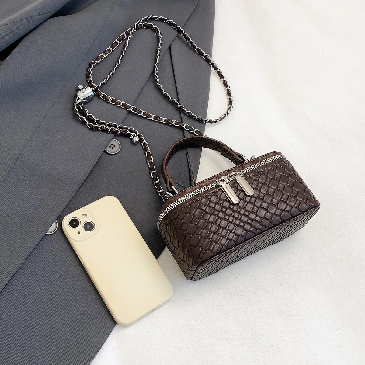 A dark brown woven PU leather crossbody bag rests next to a light-colored smartphone and a gray blazer.