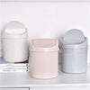 A set of three compact covered desktop trash bins in white, pink, and gray with flip lids.