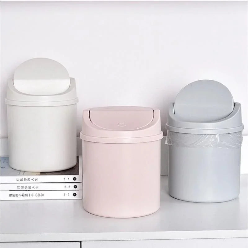 A set of three compact covered desktop trash bins in white, pink, and gray with flip lids.