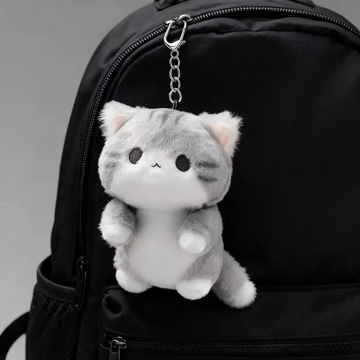 A soft grey cat plush charm hangs from a zipper pull on a black backpack.