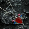 A silver pendant necklace featuring a red stone set in blue and pink accents, resting on a dark gray rock.