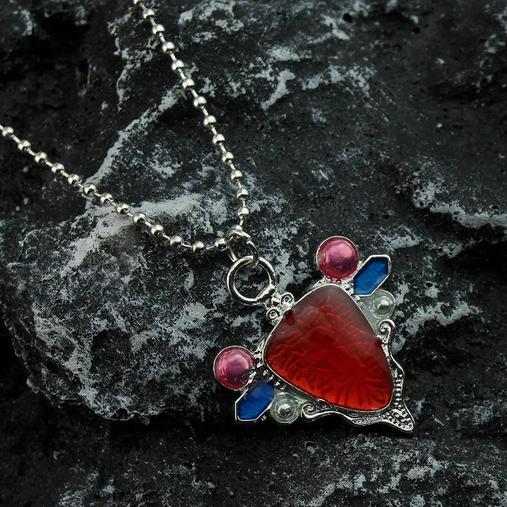 A silver pendant necklace featuring a red stone set in blue and pink accents, resting on a dark gray rock.