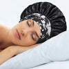 A woman is sleeping wearing a black silk satin sleep bonnet with a white floral elastic band.
