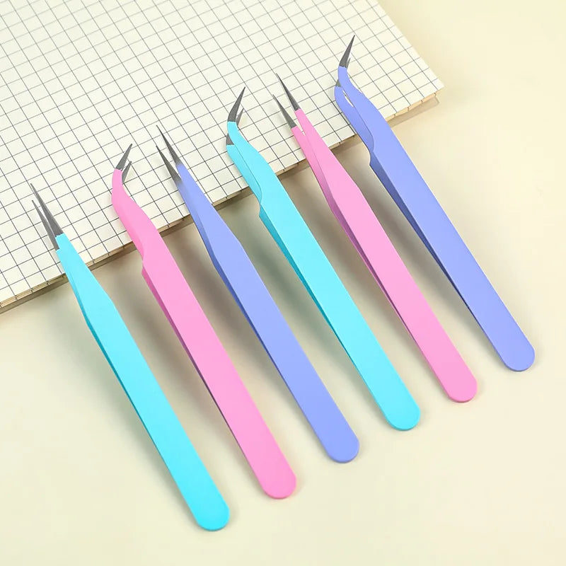 A set of five colored craft tweezers with pink, blue, and purple handles resting on a grid paper background.