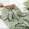 A soft sage green muslin baby swaddle blanket draped next to a woven basket.