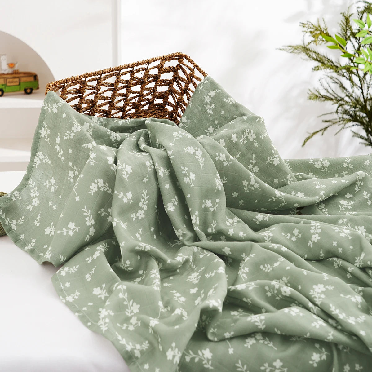 A soft sage green muslin baby swaddle blanket draped next to a woven basket.