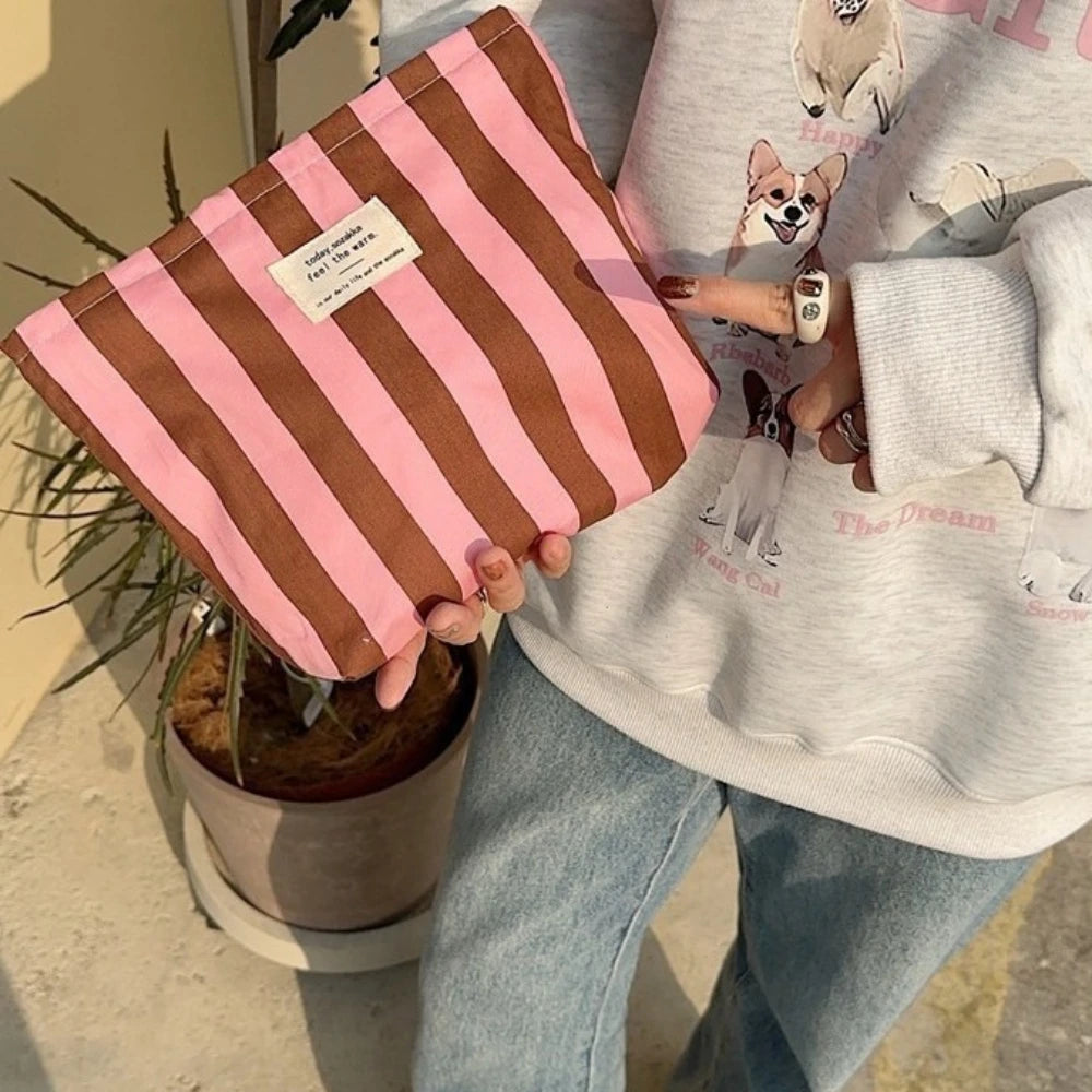 A pink and brown striped canvas cosmetic bag is held by a person wearing a light gray hoodie.