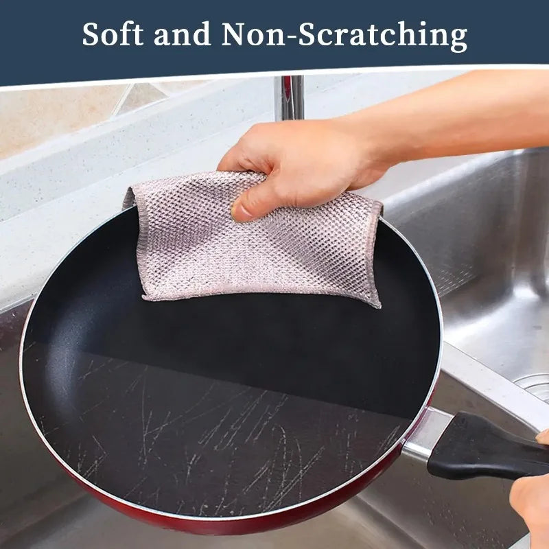 A hand is using a soft, non-scratching kitchen cleaning cloth to wipe a black frying pan in a stainless steel sink.