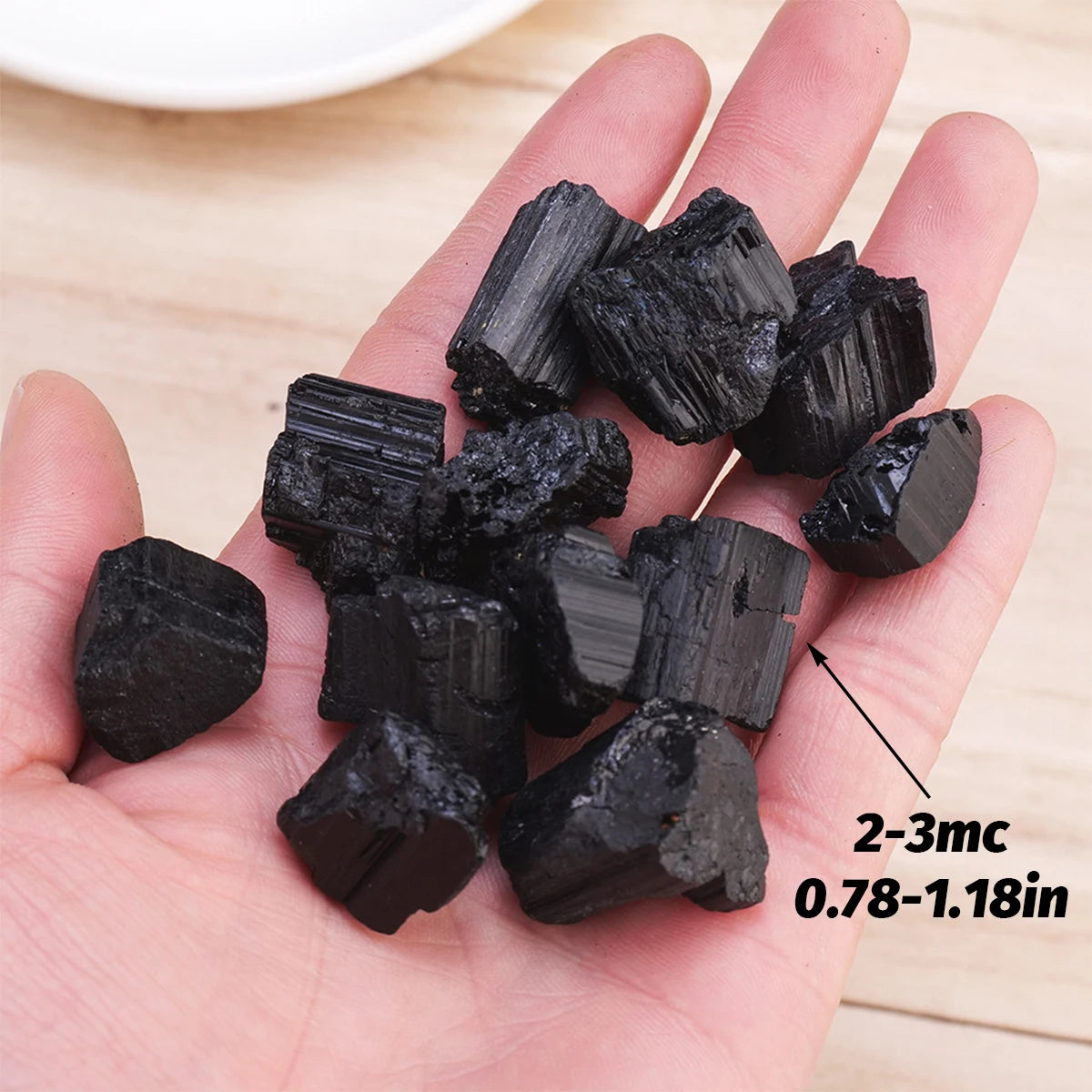 A hand holds several raw black tourmaline crystals, measuring 2-3mm in length.
