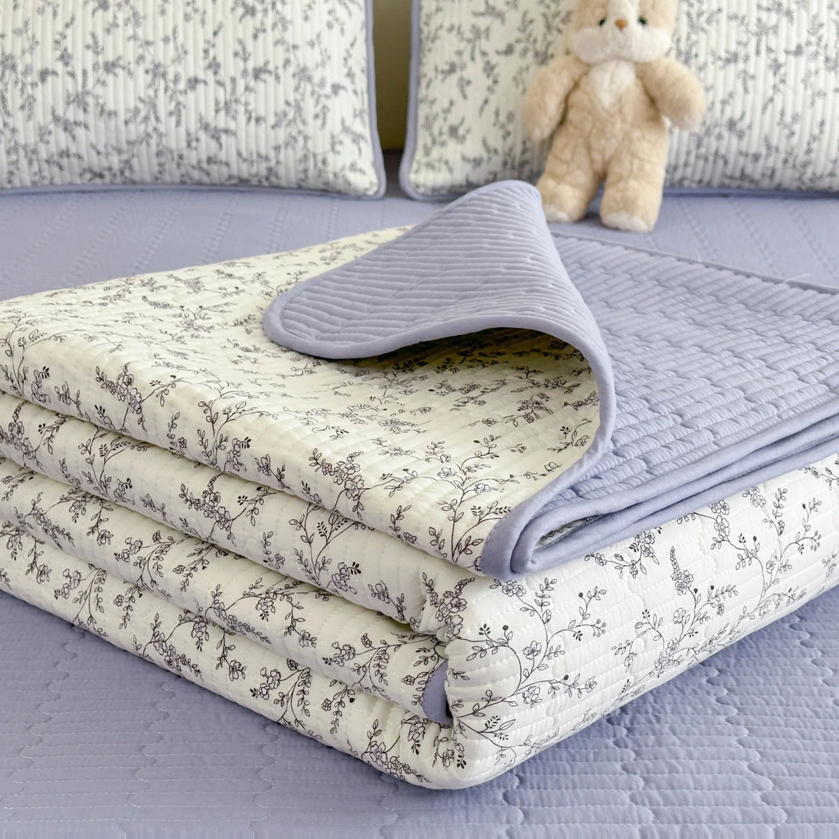 A stack of floral quilted bedspreads in white and gray patterns resting on a light purple surface.
