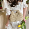 A woman holds a beige woven straw chain crossbody handbag while wearing a white dress.