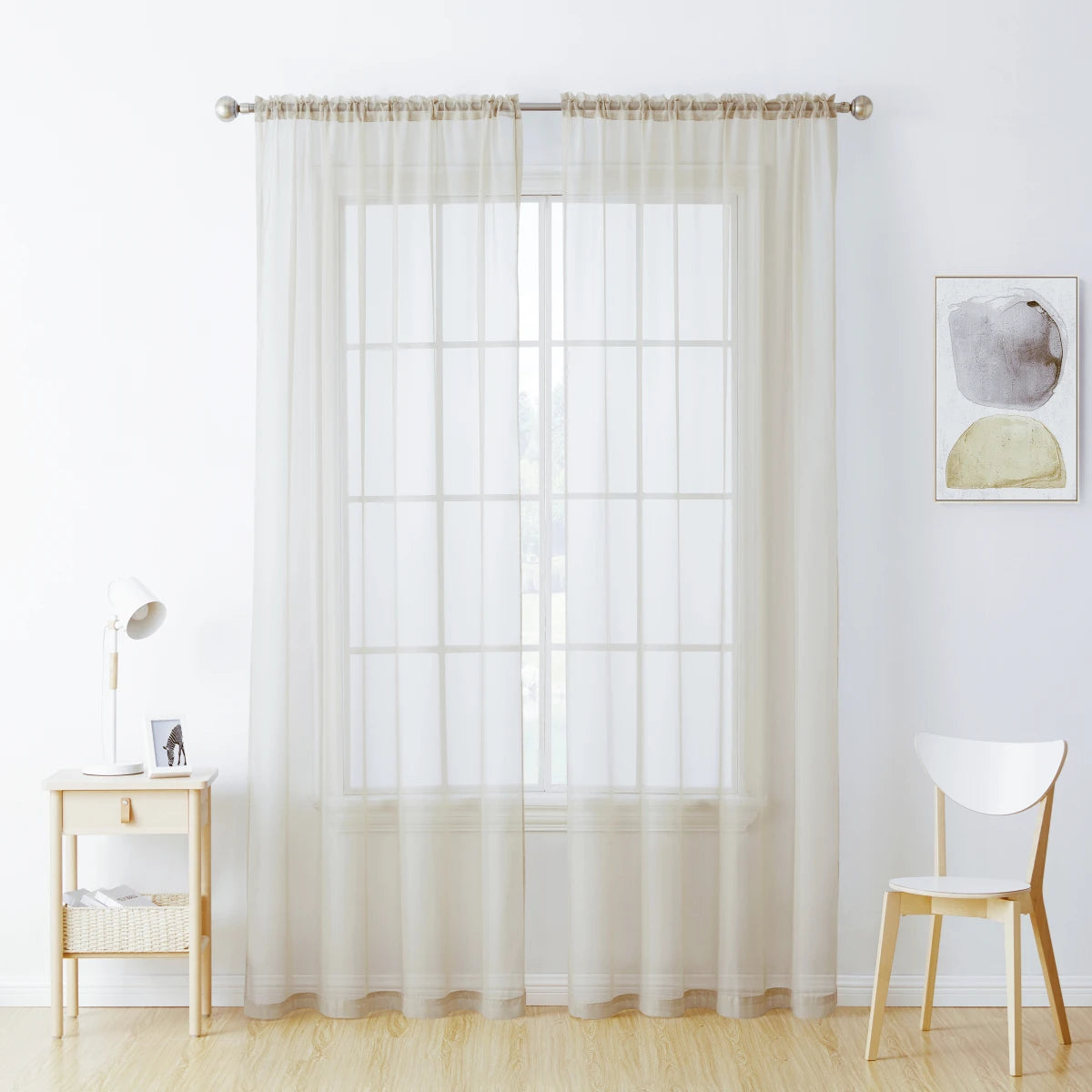 Sheer white voile curtains hanging in a window with a light wood table and chair nearby.
