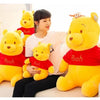 A woman is sitting on a couch with three yellow Winnie the Pooh plush toys, including one wearing a red shirt.