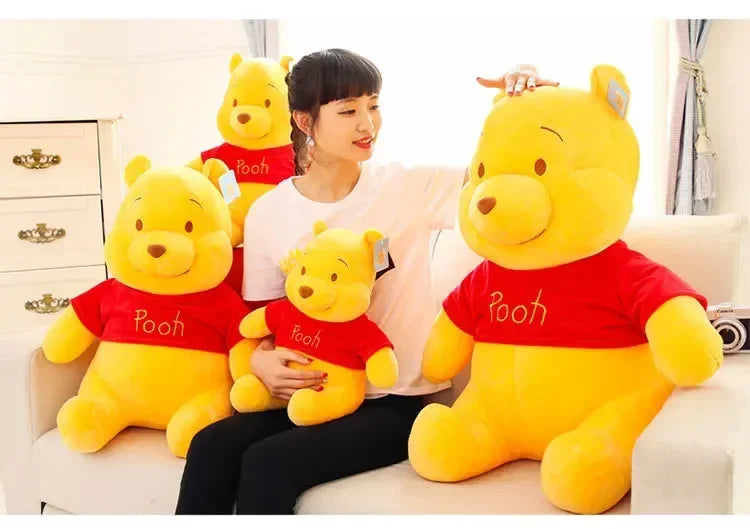 A woman is sitting on a couch with three yellow Winnie the Pooh plush toys, including one wearing a red shirt.