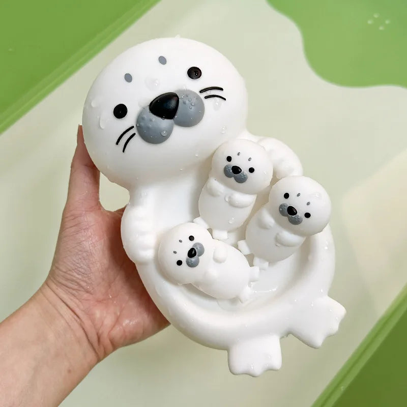 A hand holds a white silicone floating bath toy set featuring a sea otter and several small baby sea otters.