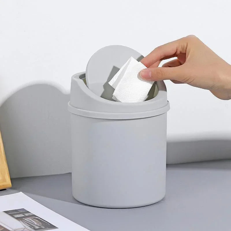 A hand is lifting a white tissue from the flip lid of a gray compact desktop trash bin.