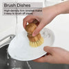 A hand is using a bamboo dish brush with natural sisal bristles to wash dishes in a sink.