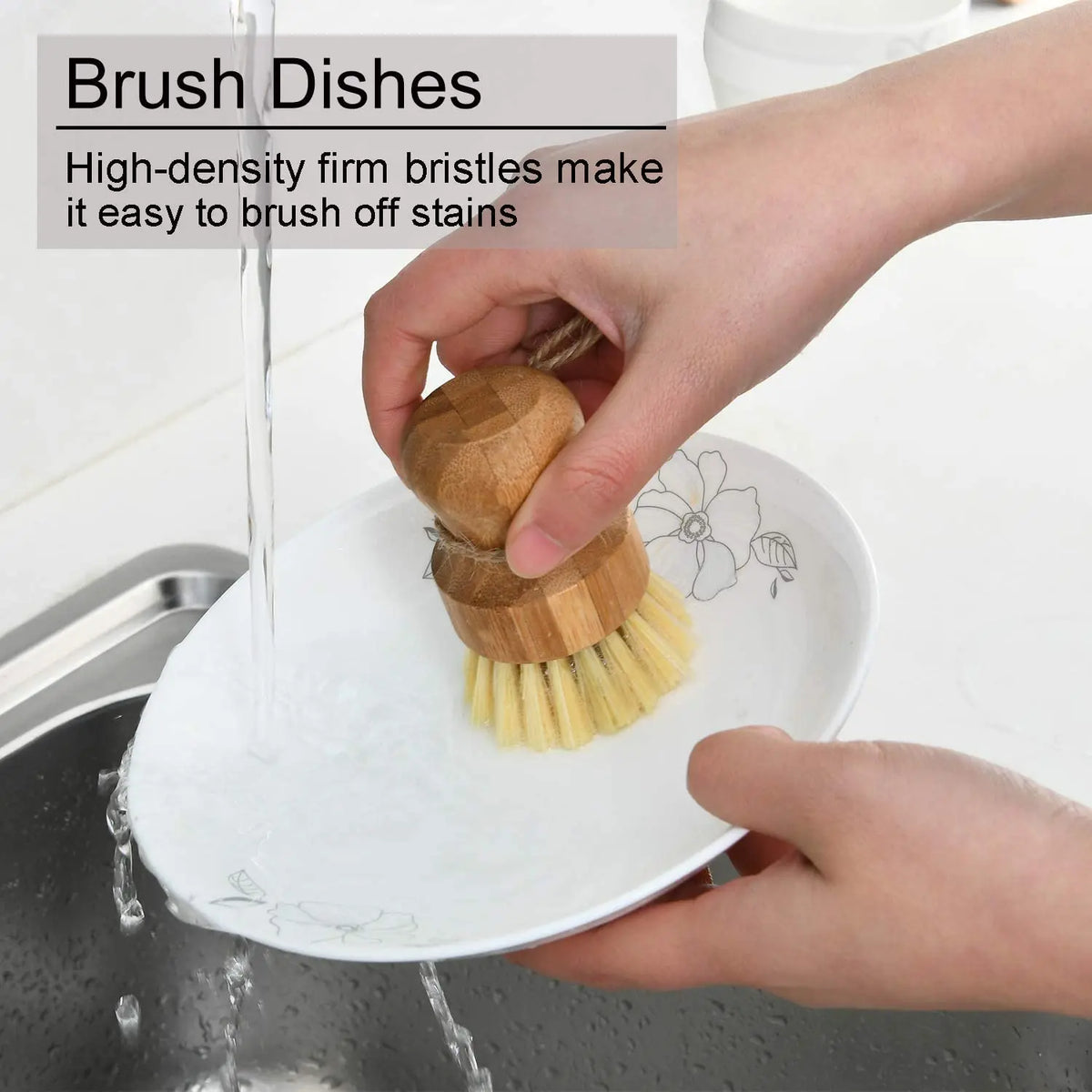 A hand is using a bamboo dish brush with natural sisal bristles to wash dishes in a sink.
