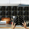 Hands holding a black marble peel-and-stick tile pattern against a dark tiled backsplash.